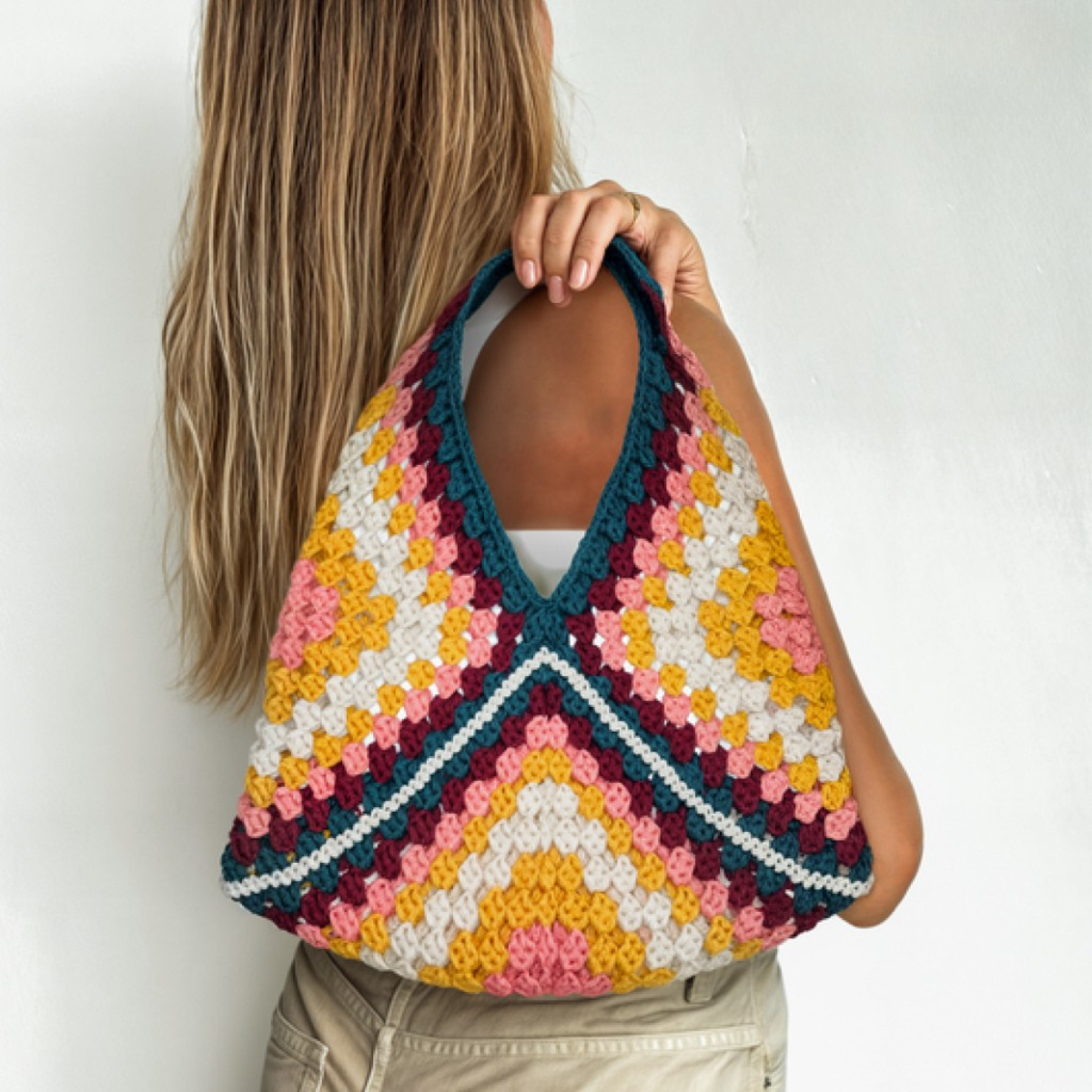 A woman with long hair holds a colorful, handmade bag made from the "6 Crochet Patterns for Must-Have Bags," featuring geometric granny square designs in pink, yellow, teal, and white over her shoulder.