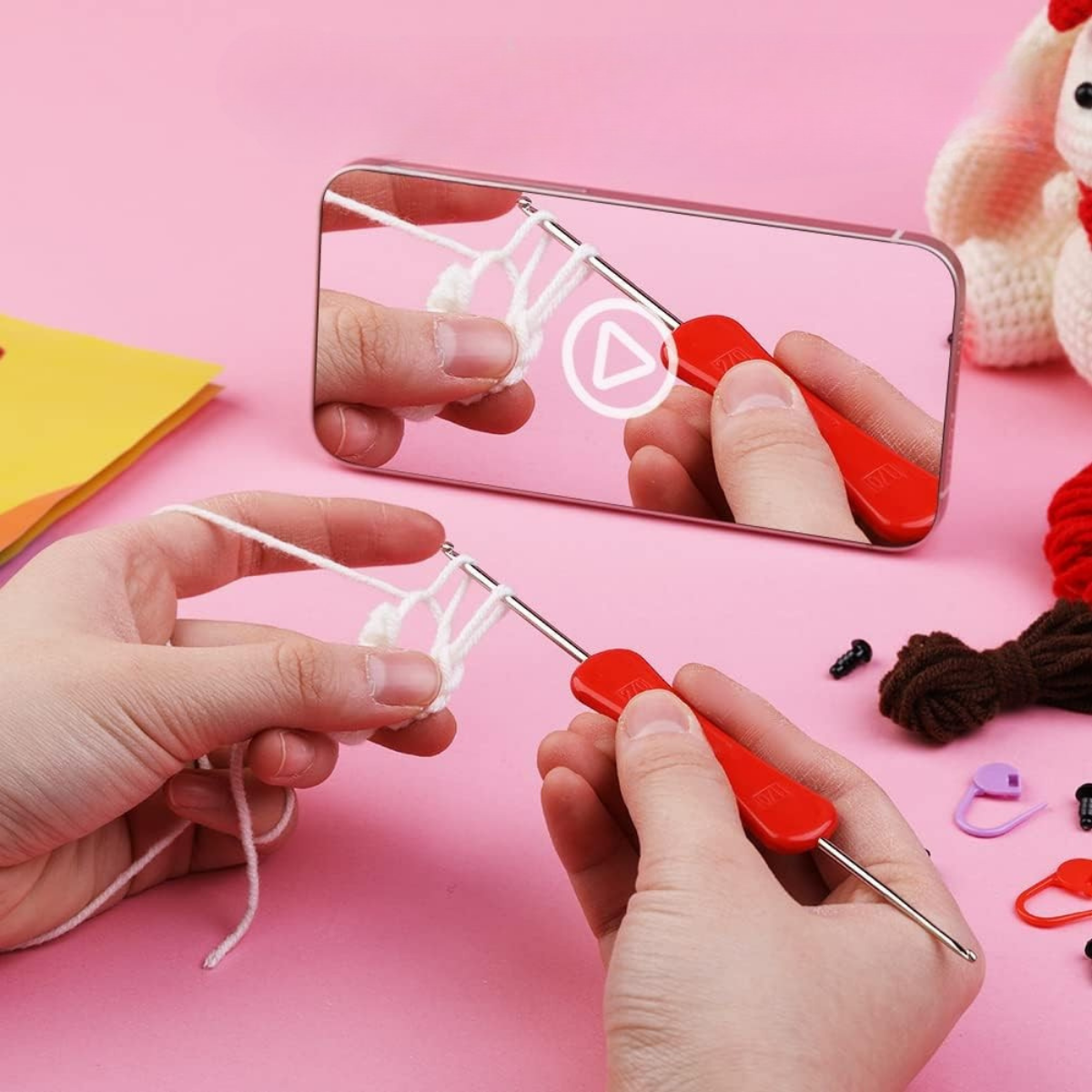 A person uses the Crochet Rabbit Kit with a red-handled crochet hook and white yarn, following a tutorial on their smartphone; craft supplies are scattered on the pink surface.