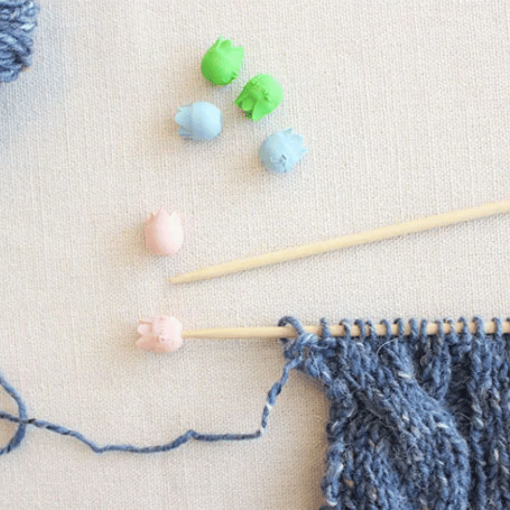 A knitting project using blue yarn on a needle is enhanced by the colorful and durable Tulip Rubber Point Protectors Small, placed on a light canvas surface to ensure needle protection.