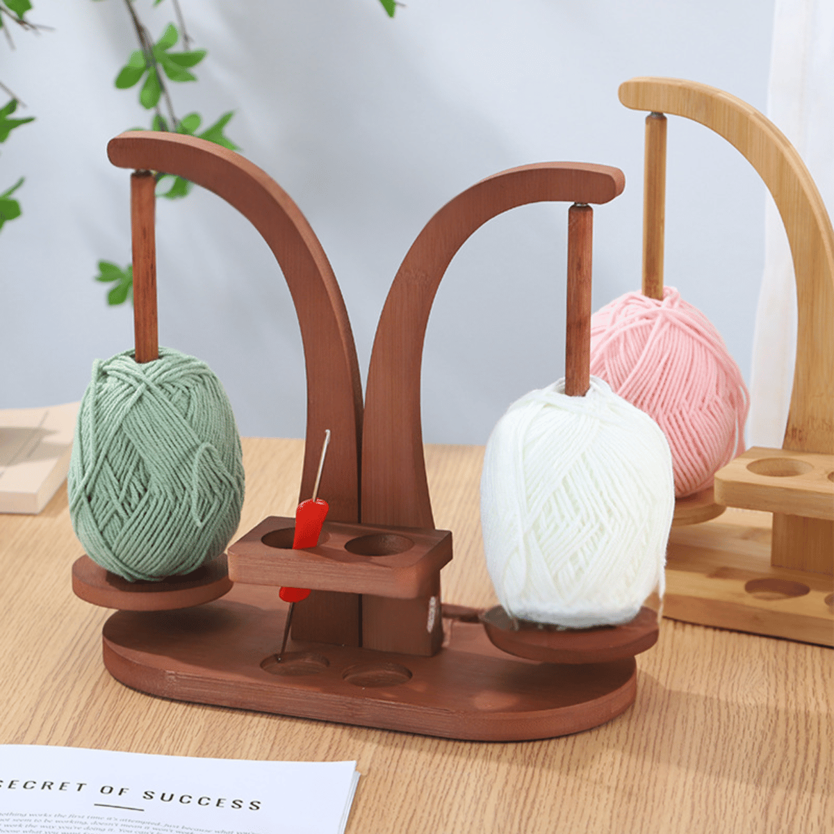 The Double Yarn Spinner Stand with Tool Storage keeps green and white yarn balls tangle-free, holds a red crochet hook, and sits on a table beside another yarn storage holder with pink yarn in the background.