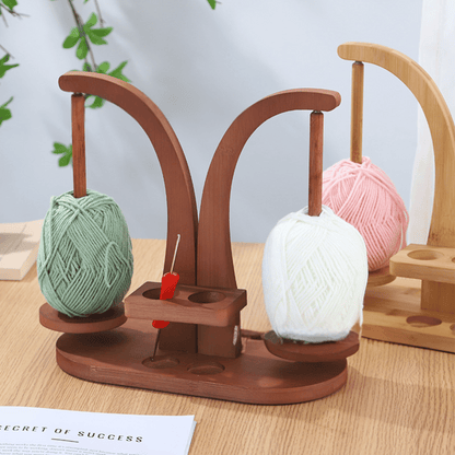 The Double Yarn Spinner Stand with Tool Storage keeps green and white yarn balls tangle-free, holds a red crochet hook, and sits on a table beside another yarn storage holder with pink yarn in the background.