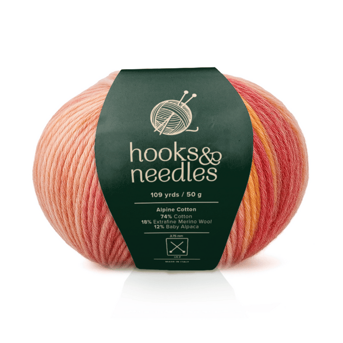 A bulky ball of Alpine Cotton yarn in a pink and orange gradient, with a green "hooks & needles" label detailing its cotton-viscose blend and yardage.