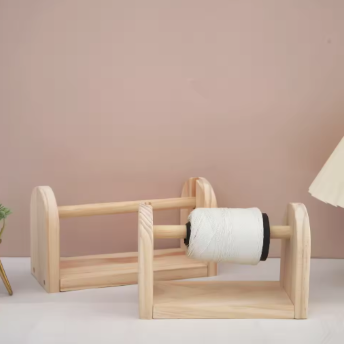 Two Wooden Yarn Dispensers are on a table—one holds white string for crafting, the other is empty. Their design enables smooth rotation, set against a simple muted pink wall.