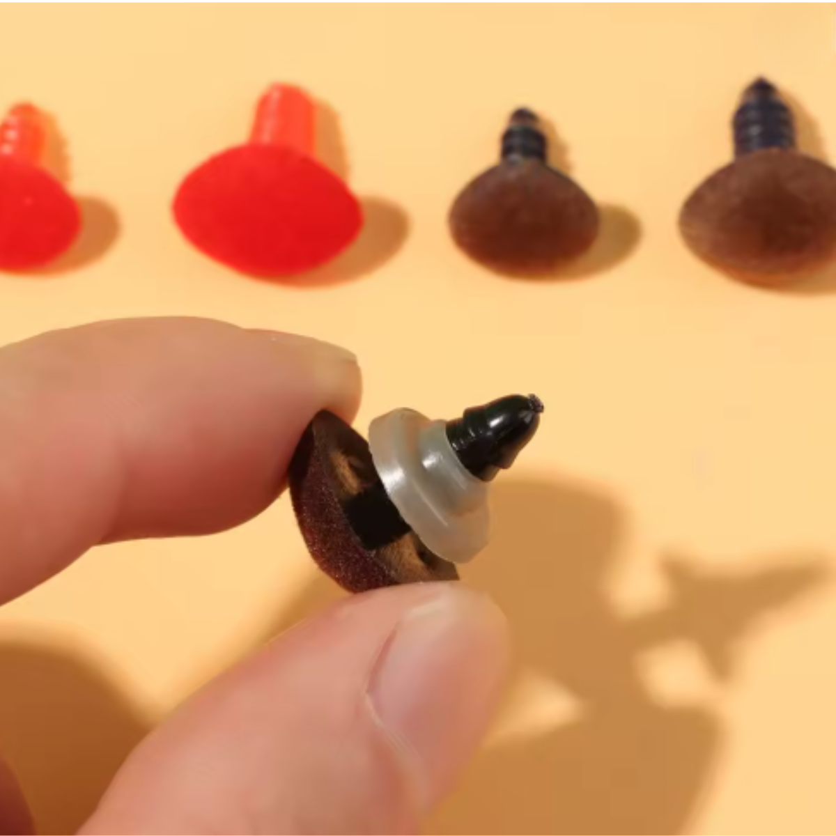 A hand holds a brown Safety Nose for Toys with Washer, while similar safety noses in red and brown are lined up on a beige background—ideal for crochet toys and amigurumi creations.