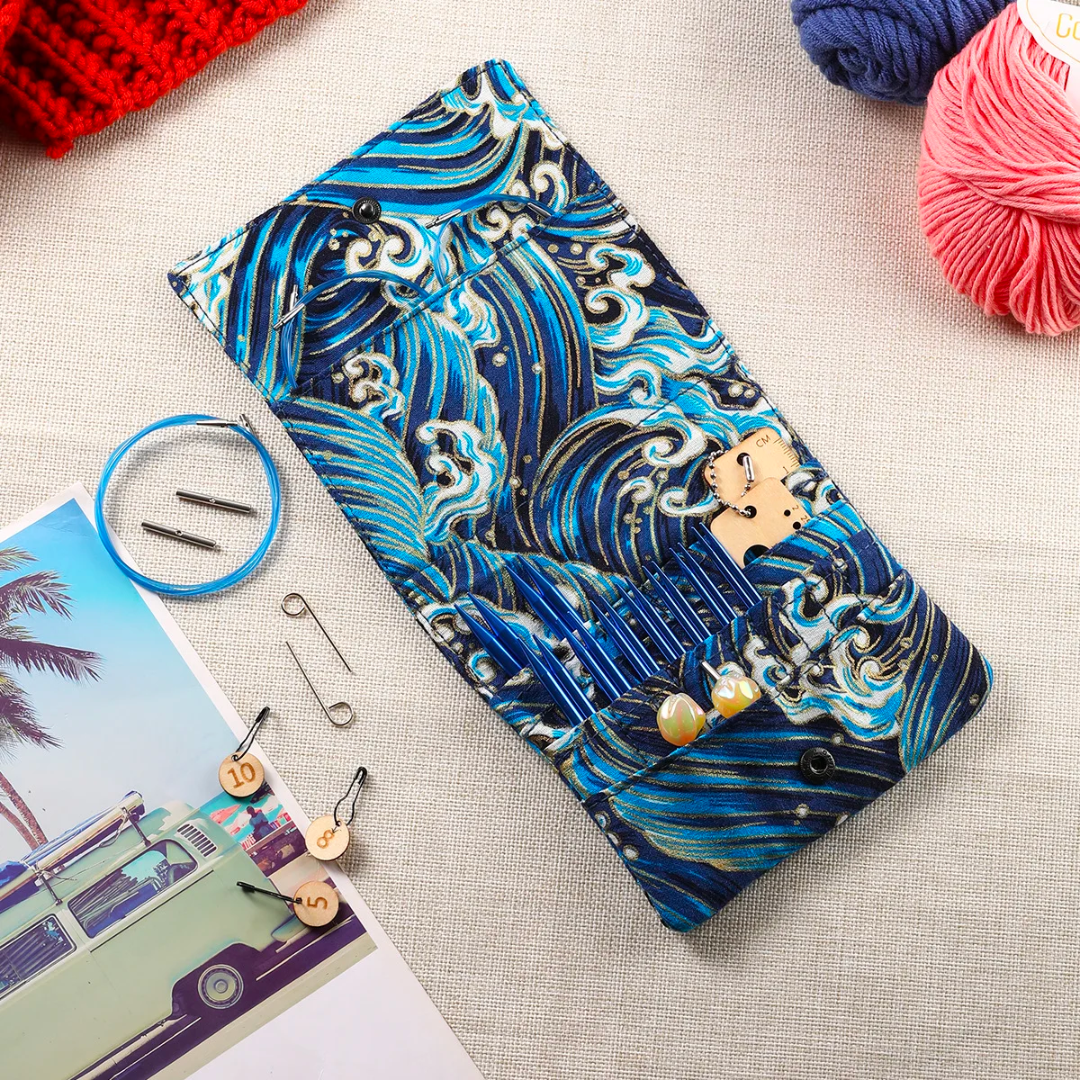 A blue wave-patterned eyeglass case on a fabric surface surrounded by Premium Aluminium Knitting Needle Set, safety pins, and a postcard with a palm tree and a vintage van.