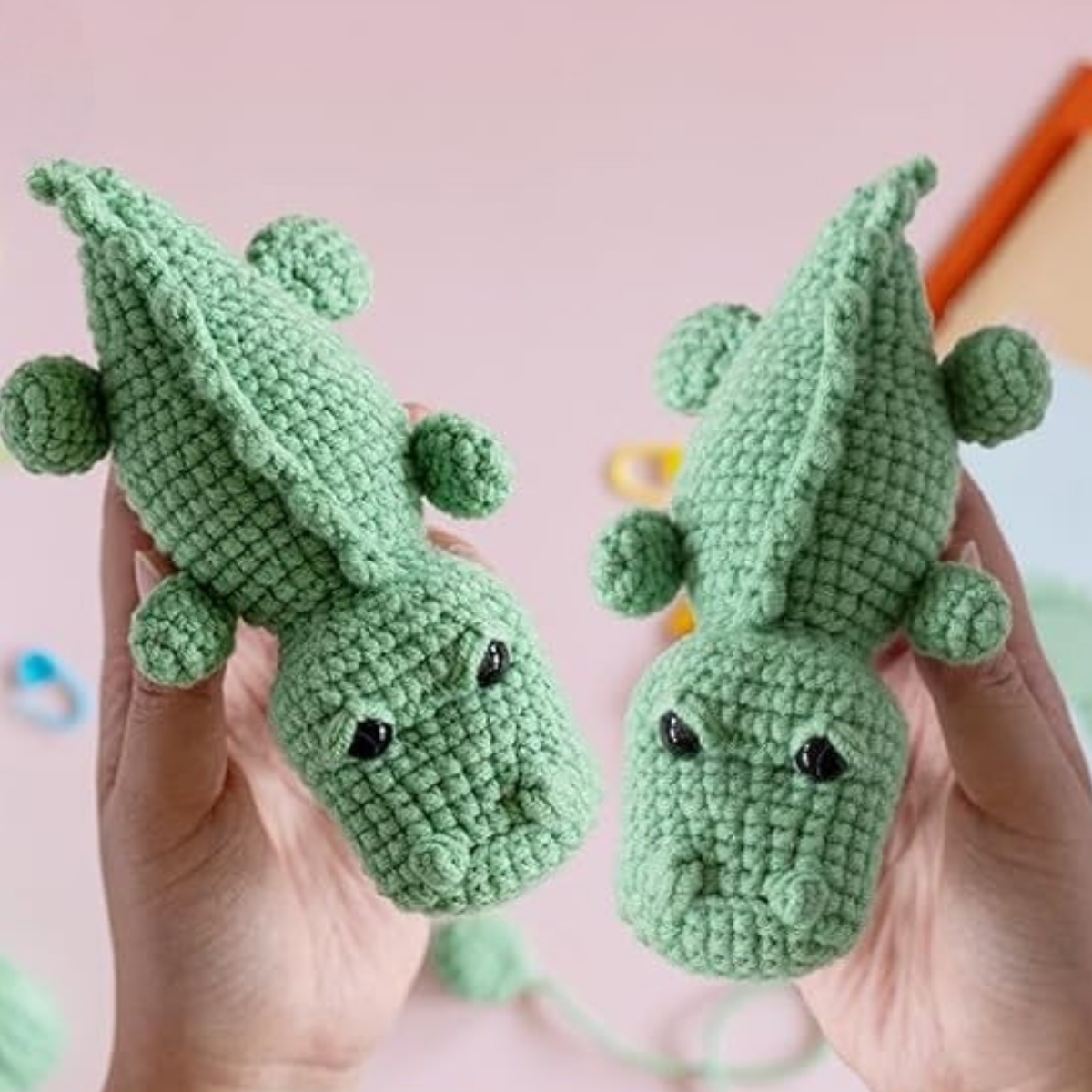 Two hands hold crocheted green crocodile toys with black eyes against a pink background scattered with crafting materials—an ideal beginner project or gift made using the Crochet Crocodile Kit 2pcs.