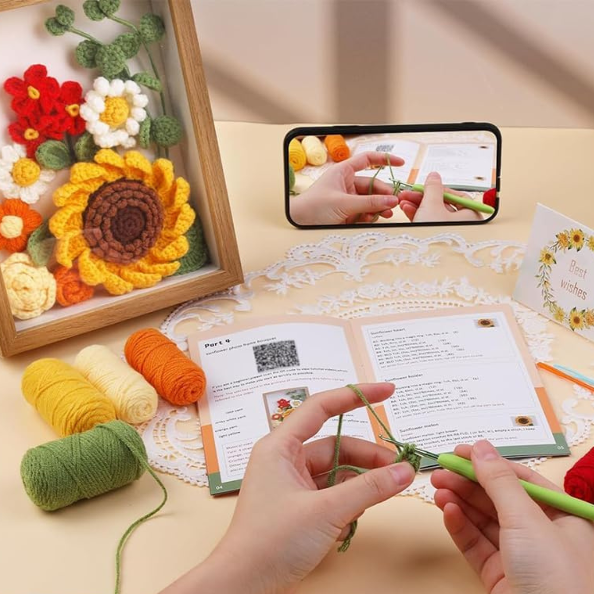 Hands crocheting with a green hook, following a pattern from the booklet; colorful yarn, a phone showing a crochet for beginners tutorial, and the Sunflower Crochet Kit with Frame—perfect for home decor—are on the table.