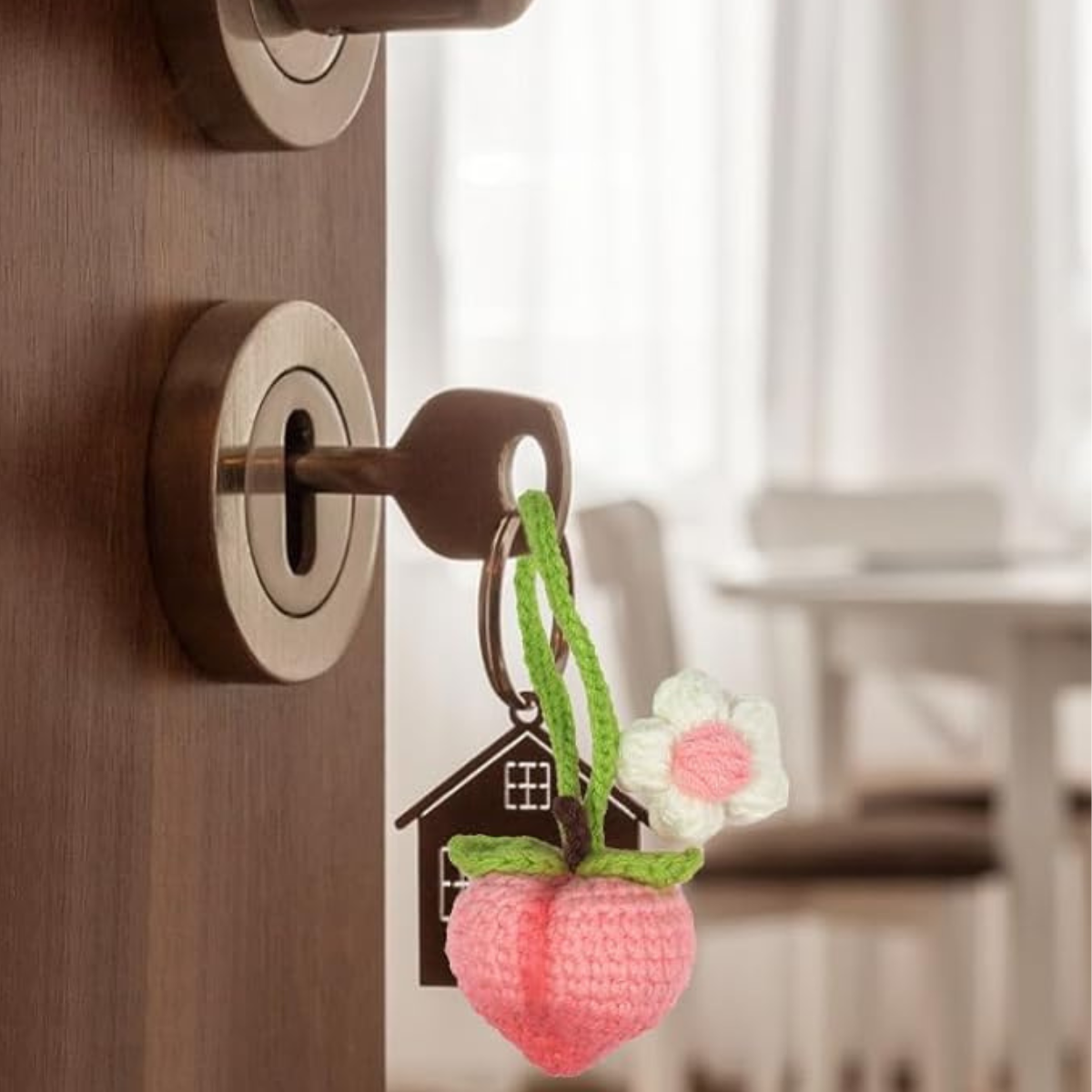 A key with a house-shaped keychain and charms from the Crochet Fruits Kit 4pcs, including a crocheted peach and flower, is inserted in a door lock, with a blurred dining area visible in the background.