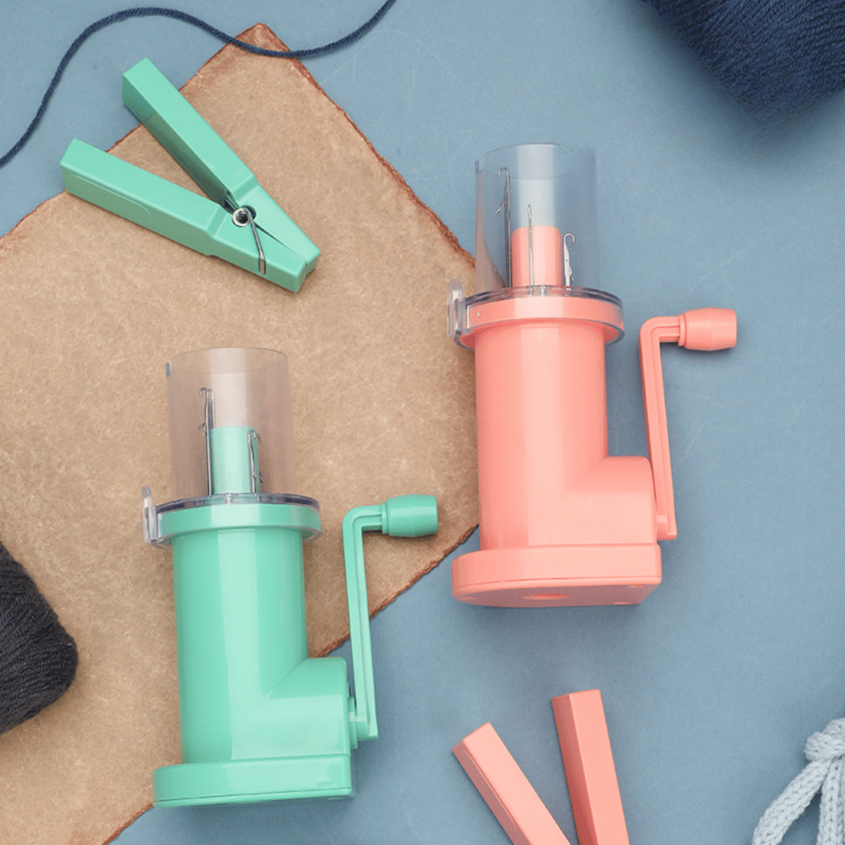 Two i-Cord Knitting Machines—mint green and peach—are displayed on a blue surface with clothespins, yarn, and brown paper. This versatile tool makes creating i-cords quick and easy.