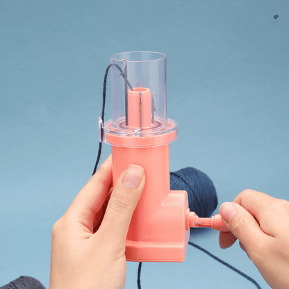 A person uses the i-Cord Knitting Machine, turning its handle to feed dark yarn through the pink device against a blue background—a quick and easy tool for making I-cords.