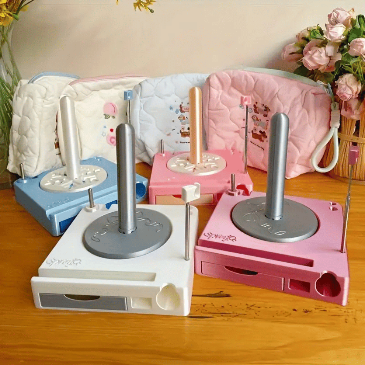 The Spinning Yarn Holder with Tool Storage comes in blue, white, pink, and rose pink, displayed on a wooden table beside three patterned pouches and a vase of flowers in the background.