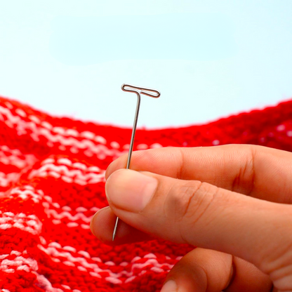 A hand holding a rust-resistant silver T-Pins for Blocking against a red and white crocheted background.
