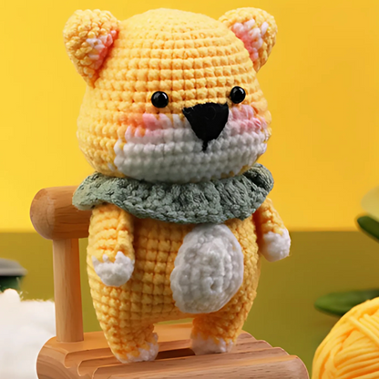 A yellow crocheted bear plush toy with a green ruffled collar, made from the Crochet Bear Kit, sits on a small wooden bench against a yellow background.