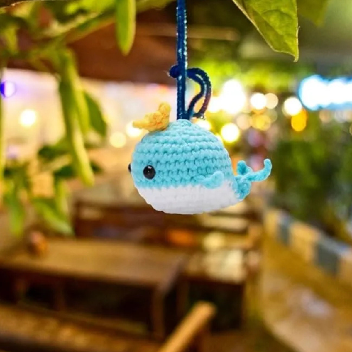 The Crochet Whale Kit 2pcs lets you make cute blue and white whale keychains, each with a tiny yellow hat—perfect for beginners. In the photo, one hangs from a branch with a softly blurred outdoor seating area in the background.