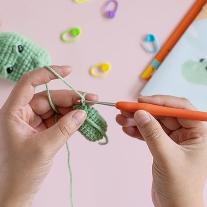 Two hands crochet with green yarn and an orange hook. Colorful stitch markers and the Crochet Crocodile Kit 2pcs, a beginner project, rest on a pink surface in the background.