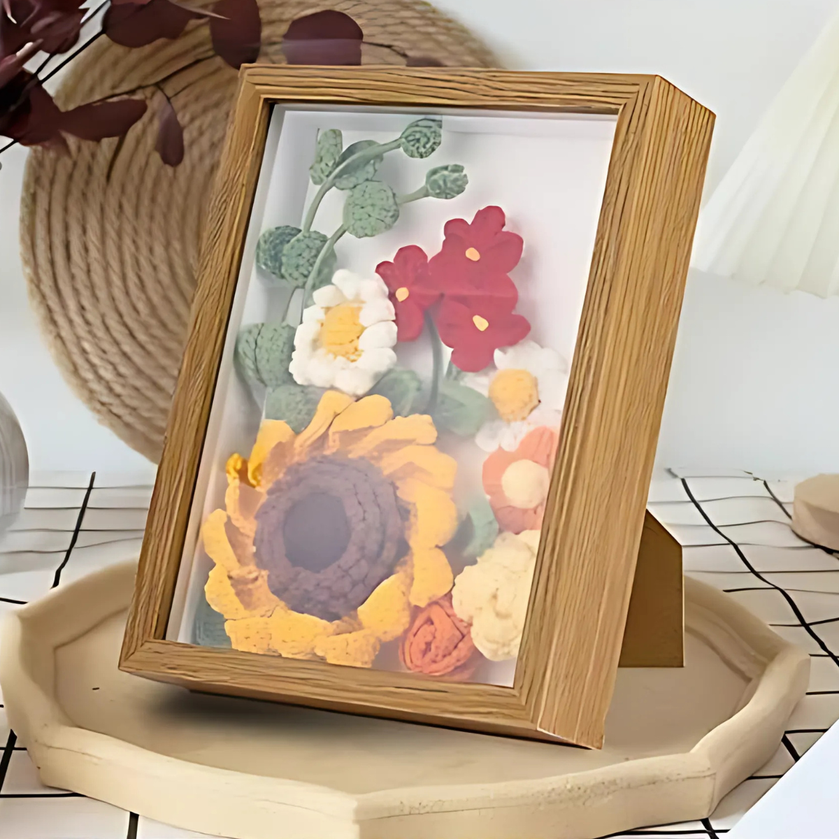 The Sunflower Crochet Kit with Frame showcases crocheted flowers in a wooden frame, displayed on a beige tray over a patterned surface—an inviting home decor accent.