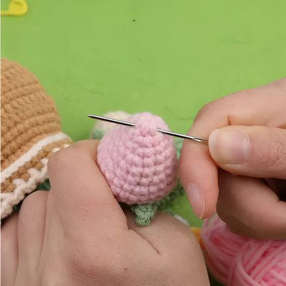 A person sews a pink piece from the Crochet Tulip Kit with a needle, holding it in one hand against a green background.