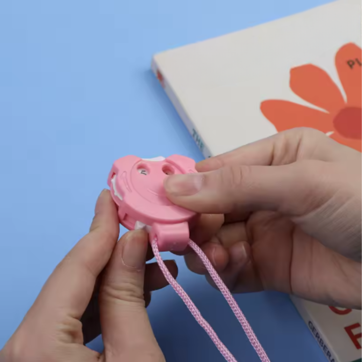 A person holds a pink Row Counter for Knitting & Crocheting with a lanyard, perfect as a crochet accessory, in front of a book featuring an orange flower on the cover.