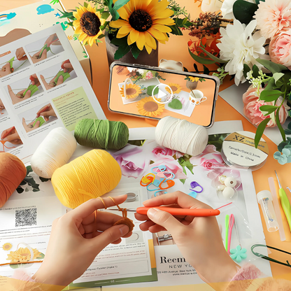 A person crochets with orange yarn at a table filled with supplies, flowers, pattern sheets, and a phone showing the Sunflower Pot Crochet Kit with 5pcs Coasters project video.