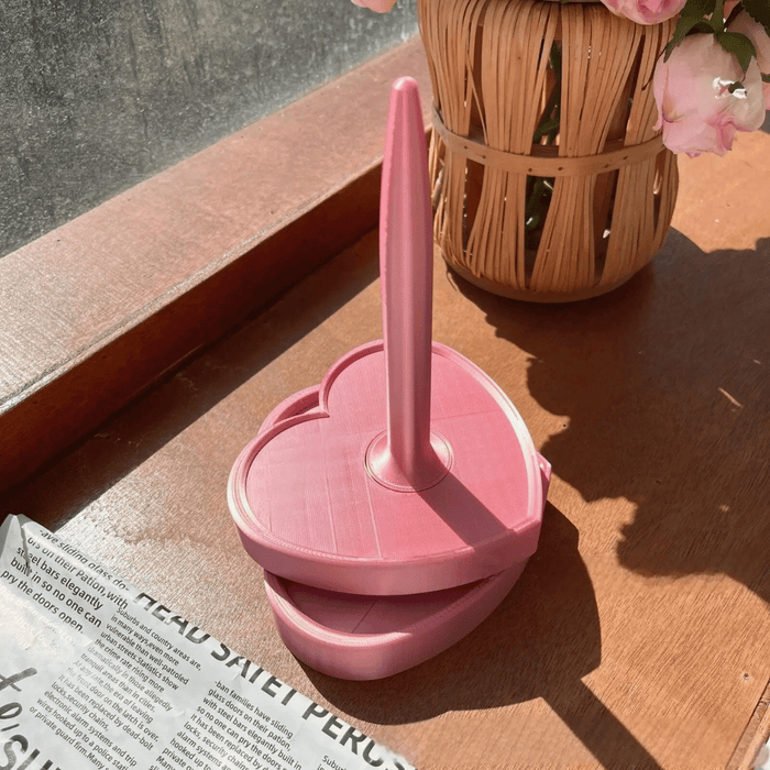 A pink, heart-shaped Spinning Yarn Holder with a vertical handle sits on a windowsill next to a woven basket of pink flowers and a newspaper.