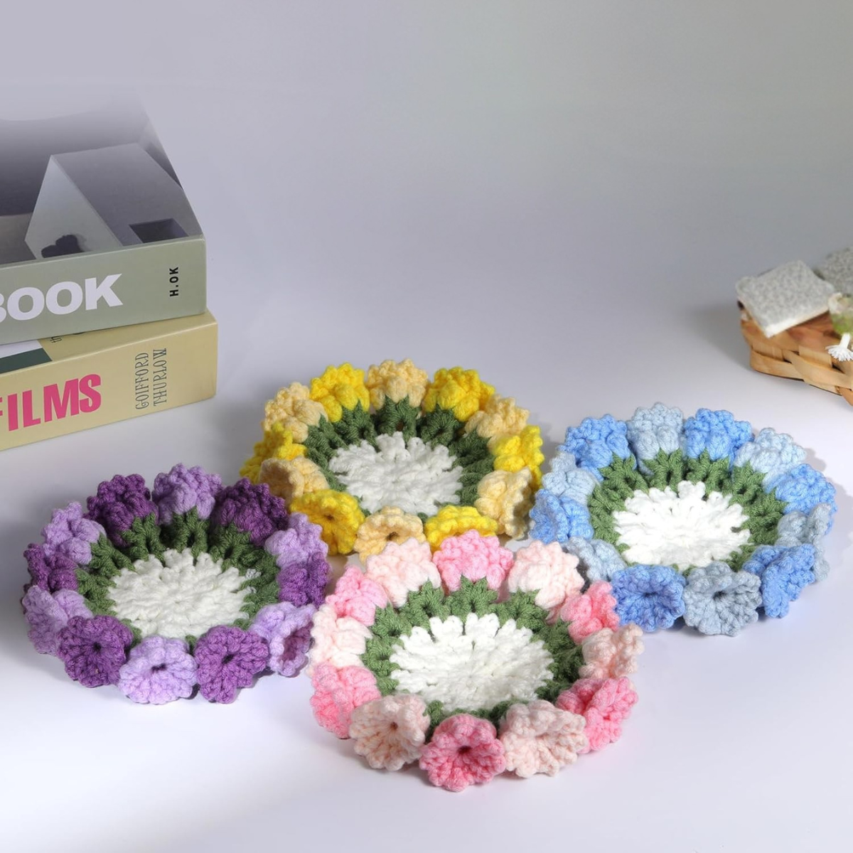 The Crochet Flower Coaster Kit 4pcs features handmade coasters with purple, yellow, blue, and pink petal designs—ideal for brightening any table. Shown on a white surface near books and a woven basket.