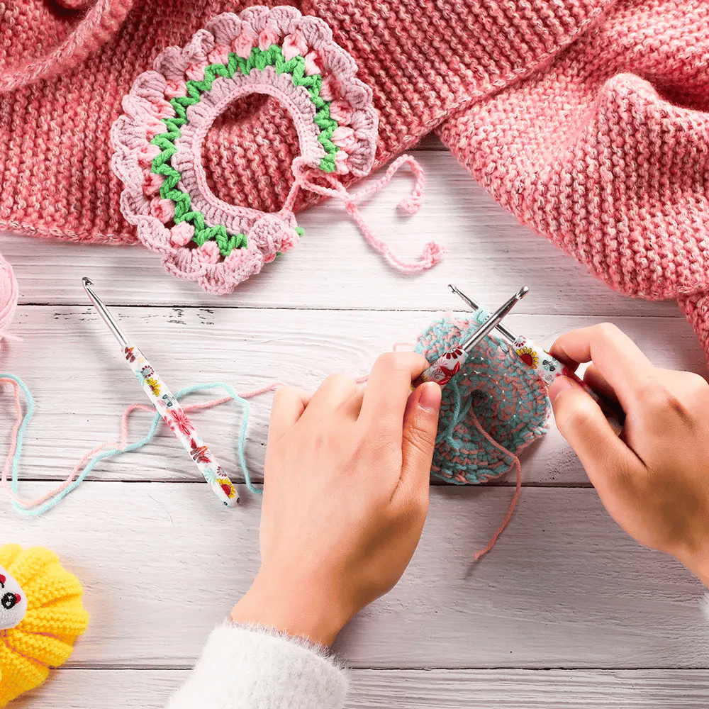 Hands knitting with colorful yarn, surrounded by crochet accessories, including a Delux Ergonomic crochet hook set, and a pink blanket on a wooden surface.
