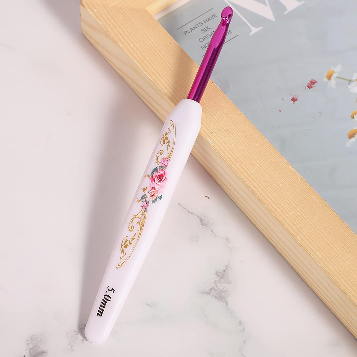 A "Crochet Hook Set 16 Pcs with Case" features a lightweight crochet hook with a white handle, pink floral designs, and metallic pink 5.0mm tip, resting on marble near a wooden frame—blending artistry and function.