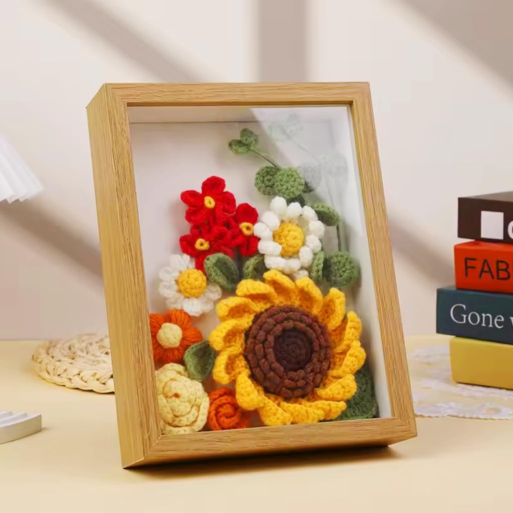 The Sunflower Crochet Kit with Frame showcases vibrant crocheted flowers—including a sunflower—in a wooden frame, set on a beige tabletop by books and a woven coaster, making charming home decor for crochet beginners.