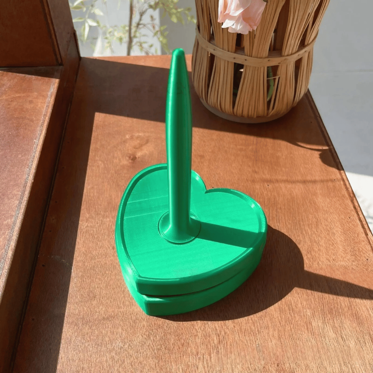A green, heart-shaped Spinning Yarn Holder with a vertical handle sits on a wooden surface in sunlight, with a wicker basket in the background.