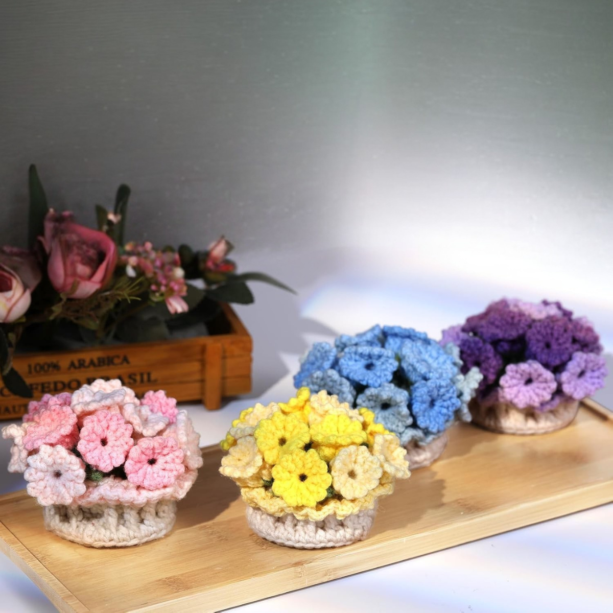 The Crochet Flower Coaster Kit 4pcs features handmade pastel pink, yellow, blue, and purple coasters with floral designs. Displayed on a wooden tray, their charm is complemented by a background box of artificial flowers.