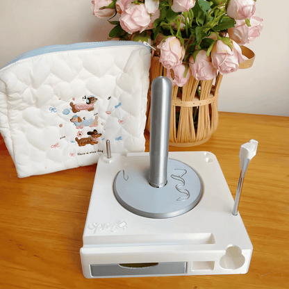 A Spinning Yarn Holder with Tool Storage featuring a silver handle and a white quilted pouch, displayed beside a basket of pink roses on a wooden surface.
