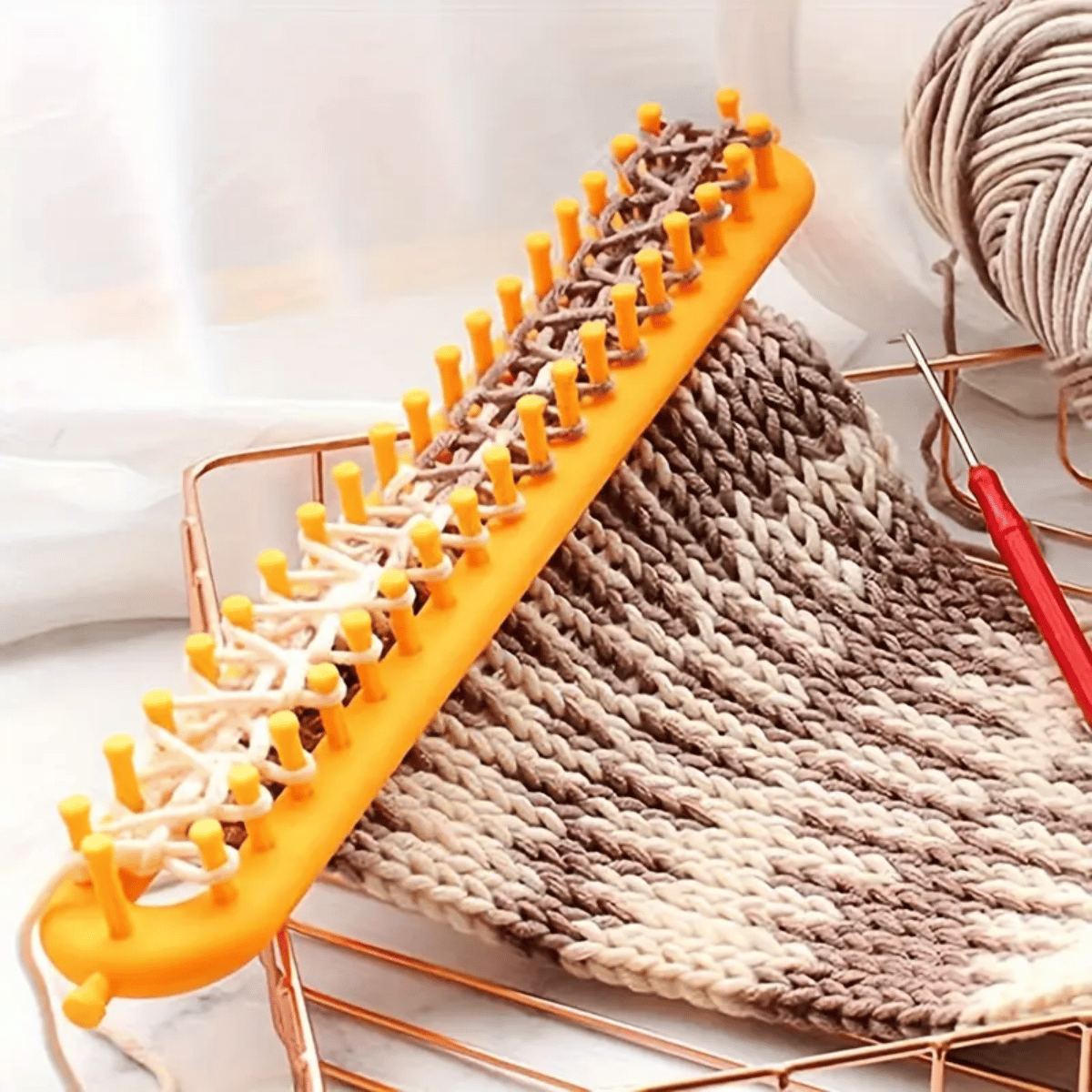 The Knitting Loom Set includes a rectangular orange loom with partially knitted beige and brown yarn, a red crochet hook, and a yarn ball—ideal for beginners and quick, efficient knitting projects.