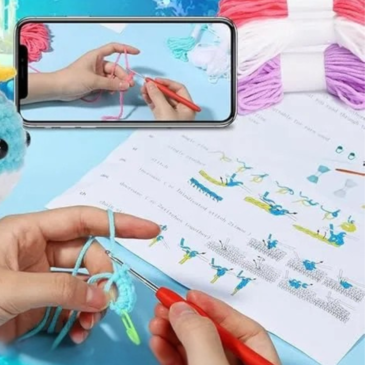 Hands crochet with blue yarn using a red hook, following beginner instructions and a video tutorial on a phone, with skeins of yarn and the Crochet Whale Kit 2pcs—including plush toy materials and whale patterns—nearby.