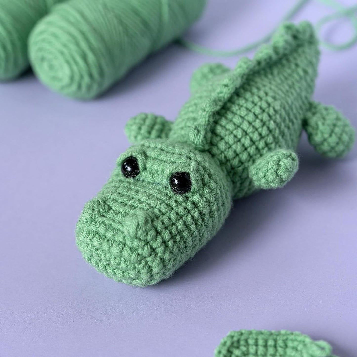 The Crochet Crocodile Kit 2pcs features a crocheted green crocodile with black bead eyes on a purple surface, with green yarn skeins in the background—ideal inspiration for your next beginner crochet project.