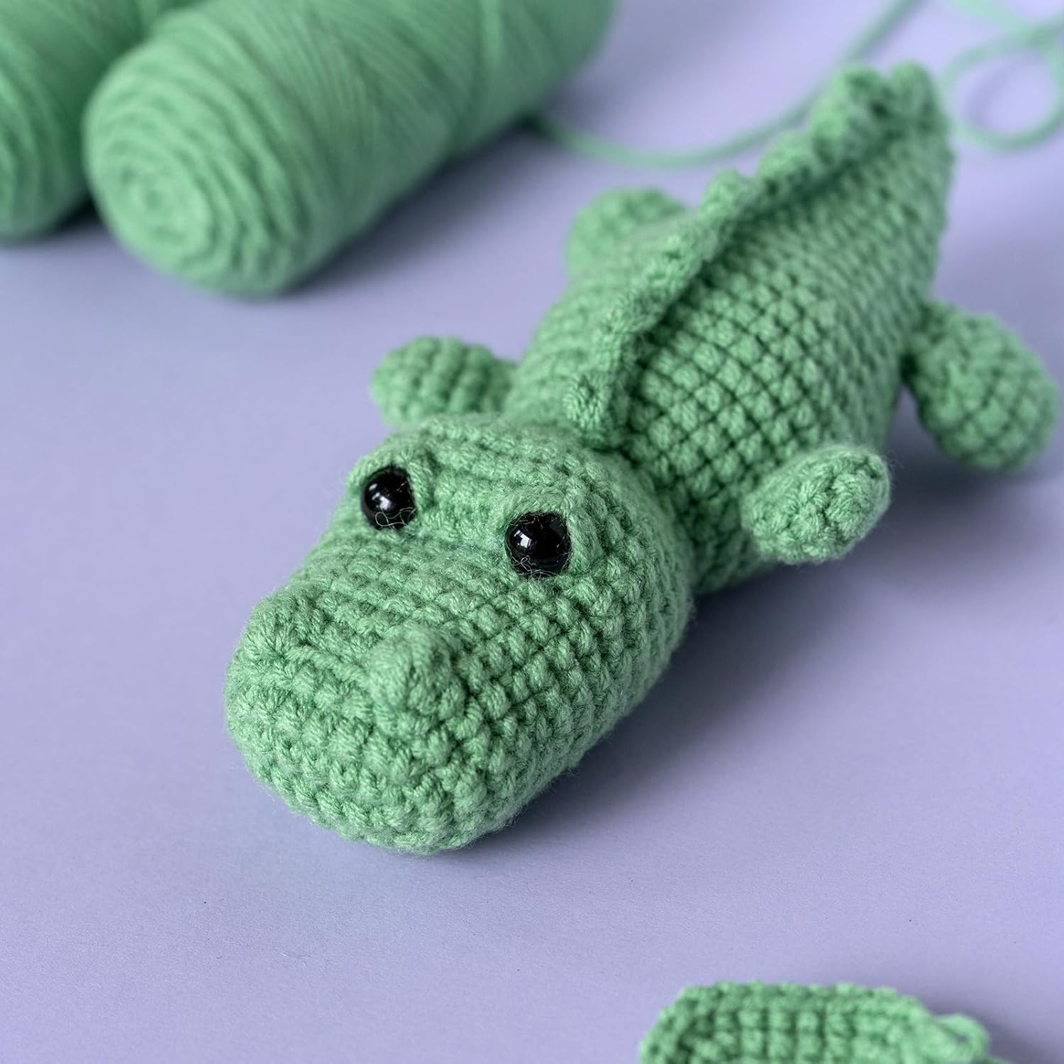 The Crochet Crocodile Kit 2pcs features a crocheted green crocodile with black bead eyes on a purple surface, with green yarn skeins in the background—ideal inspiration for your next beginner crochet project.