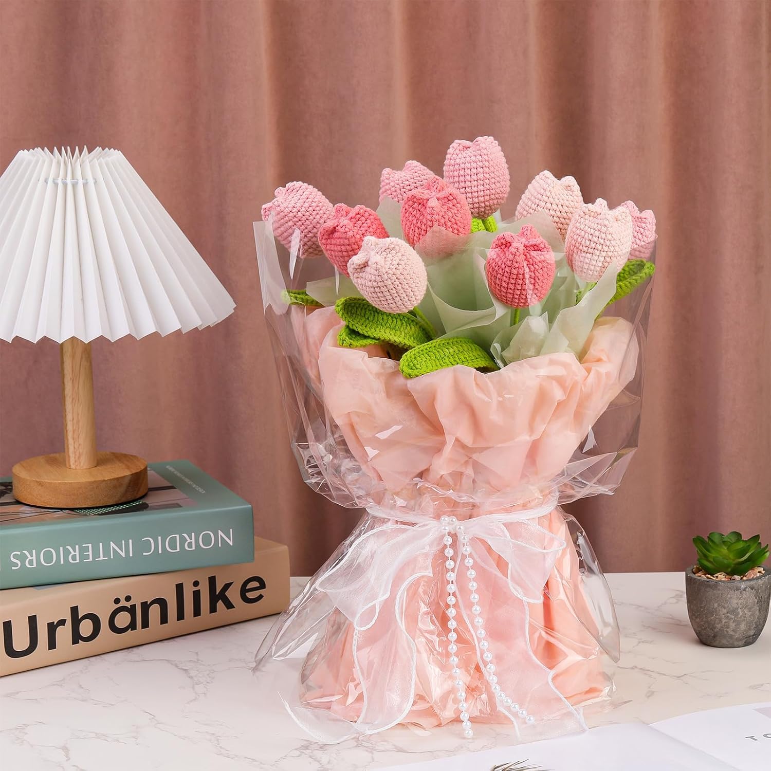 The Crochet Tulip Bouquet Kit features pink and light red everlasting tulips, wrapped in cellophane and tissue paper, beautifully displayed on a white table next to books, a lamp, and a small potted plant.