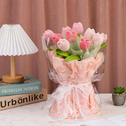 The Crochet Tulip Bouquet Kit features pink and light red everlasting tulips, wrapped in cellophane and tissue paper, beautifully displayed on a white table next to books, a lamp, and a small potted plant.