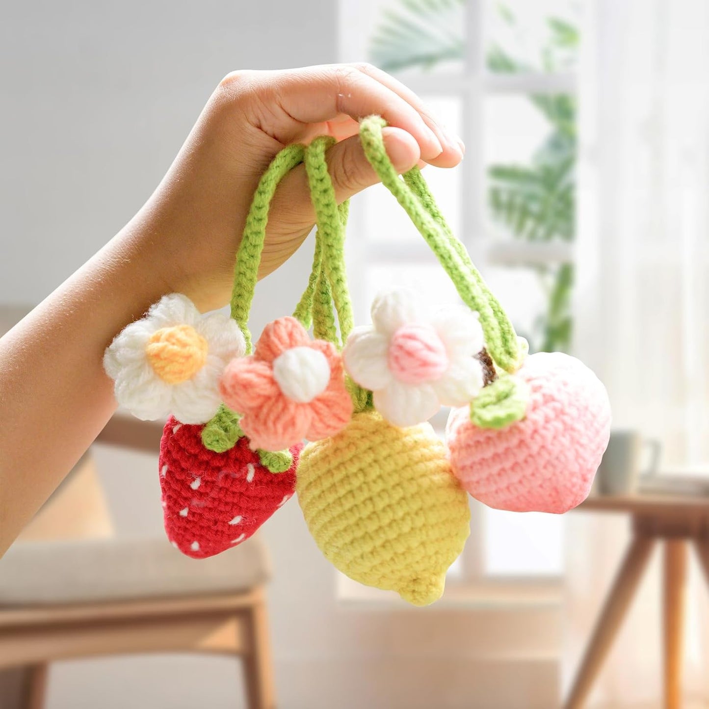 A hand displays three vibrant crochet fruit pendants—a strawberry, lemon, and peach—each with crocheted flowers. The Crochet Fruits Kit 4pcs is perfect for charming DIY projects or adding to your collection in a bright indoor setting.