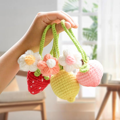 A hand displays three vibrant crochet fruit pendants—a strawberry, lemon, and peach—each with crocheted flowers. The Crochet Fruits Kit 4pcs is perfect for charming DIY projects or adding to your collection in a bright indoor setting.