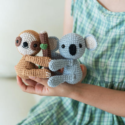 A person in a green checked dress holds two plush toys—a brown sloth and a gray koala made with the Crochet Sloth & Koala Kit—hugging a small crocheted branch.