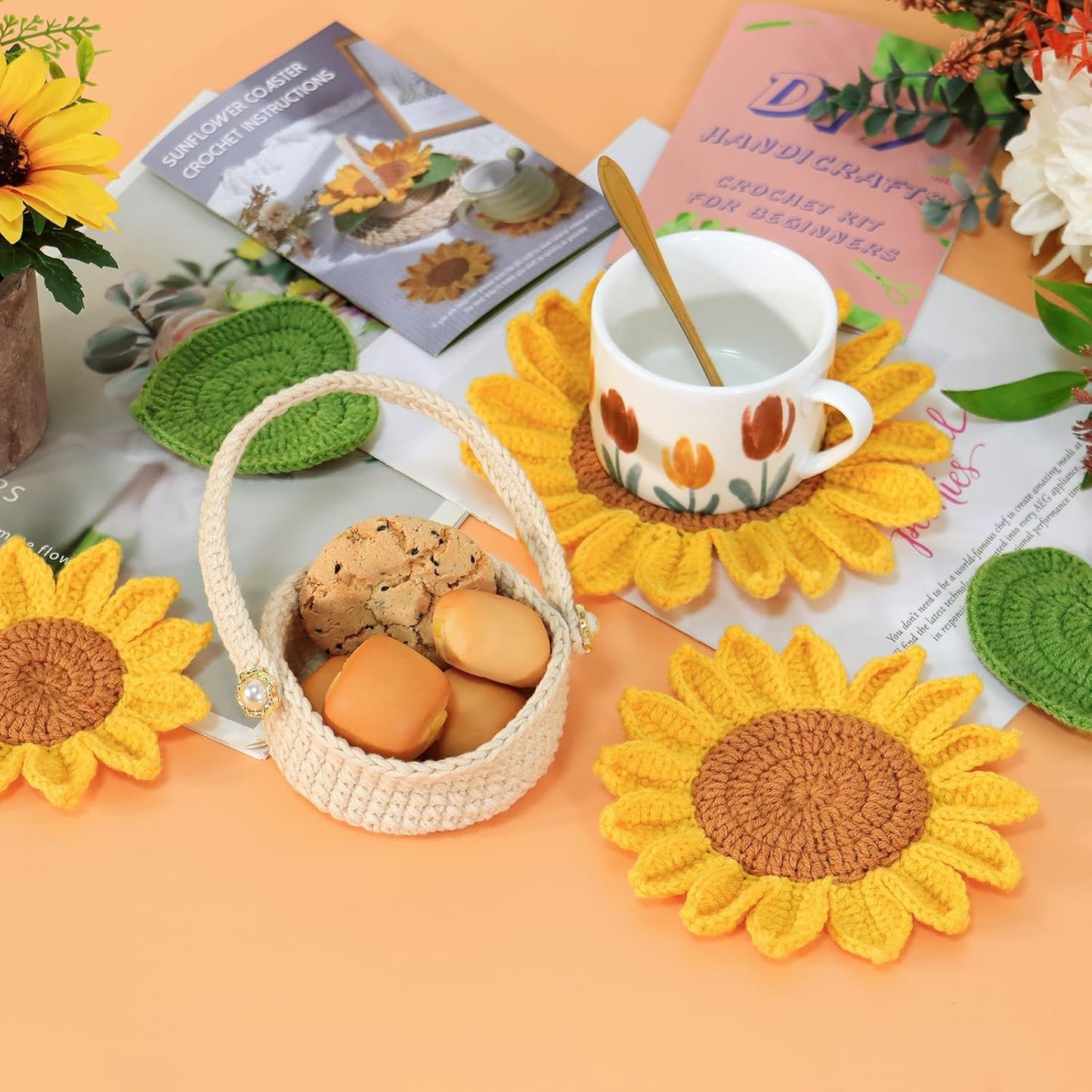 A table features the Sunflower Pot Crochet Kit with 5pcs Coasters, a cup with a spoon, cookies in a small basket, flowers, and magazines on a peach-colored surface.