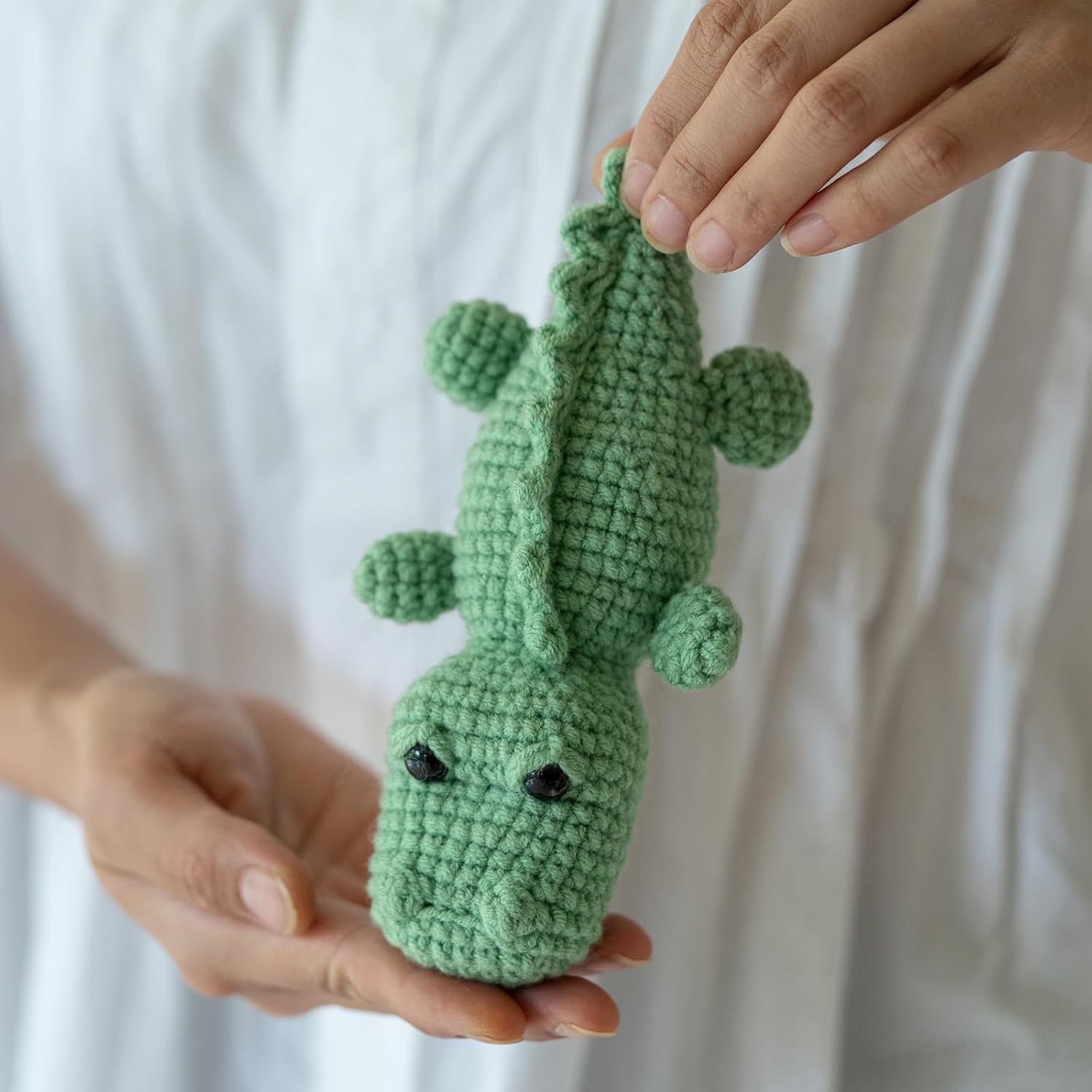 A person holds a small green crocodile toy made with the Crochet Crocodile Kit 2pcs, featuring black eyes, against a white background.