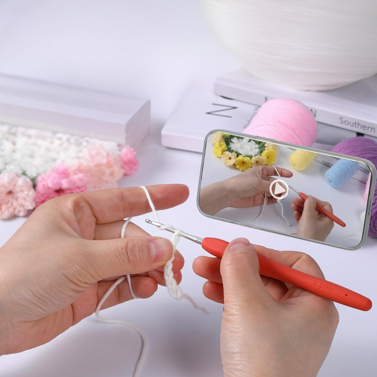 Hands crocheting with a red hook and white yarn, pastel yarn balls, books, and a phone showing a crochet tutorial—ideal for making floral coasters with the Crochet Flower Coaster Kit 4pcs.