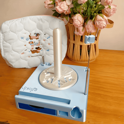 The Spinning Yarn Holder with Tool Storage, featuring a blue design, vertical spindle, and metal rods, sits on a wooden table next to pink roses and a white cushion, keeping your yarn flow smooth and organized.