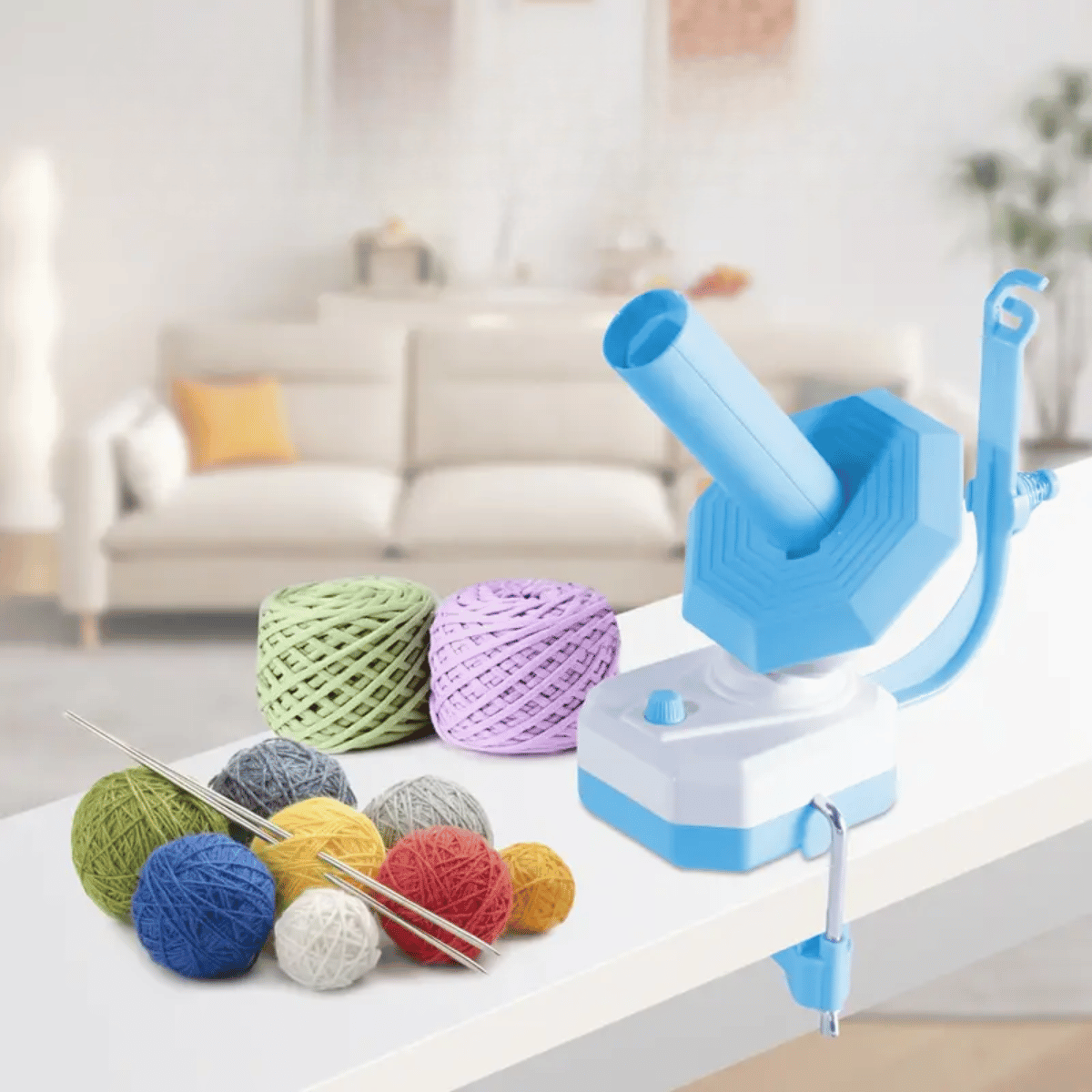 An Electric Yarn Winder, along with colorful yarn balls and knitting needles, is displayed on a table with a sofa in the background.
