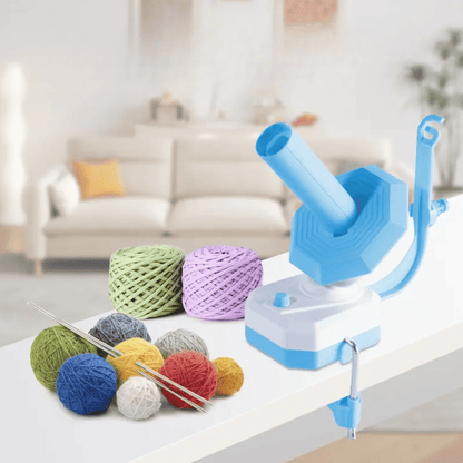 An Electric Yarn Winder, along with colorful yarn balls and knitting needles, is displayed on a table with a sofa in the background.