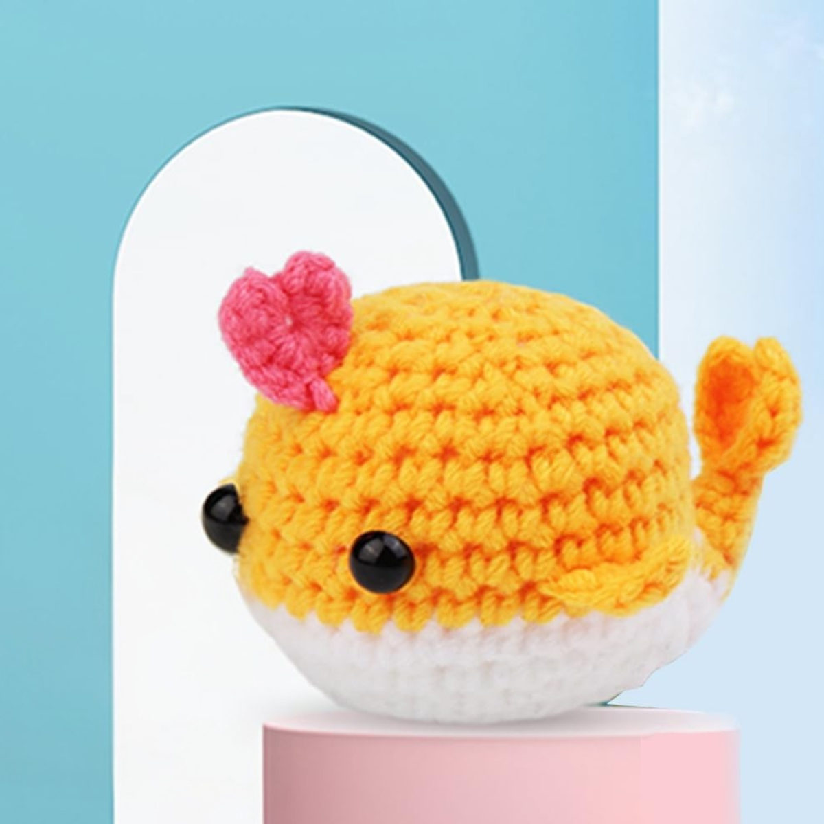 The Crochet Whale Kit 2pcs lets you make small yellow crocheted whales with white undersides, black eyes, and pink heart details, perfect for beginners. Displayed on a pastel blue and white background.