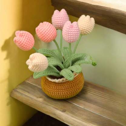 A brown crochet pot made with the Crochet Tulip Kit holds six pink and cream tulips with green stems and leaves, displayed on a wooden step beside a yellow wall.