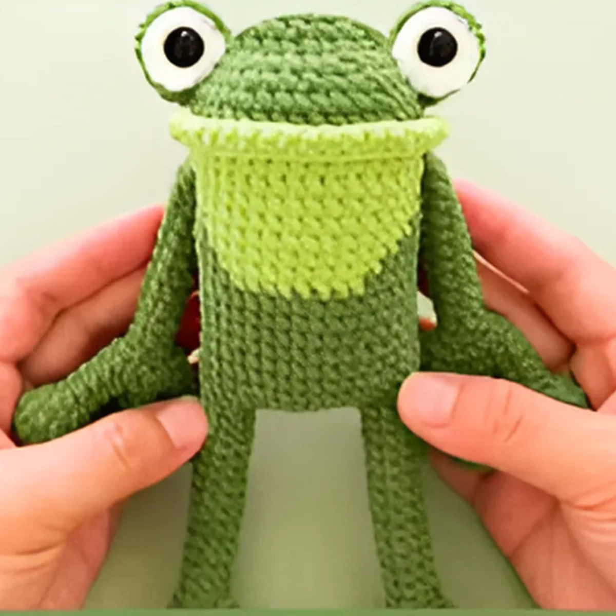 Two hands hold a green amigurumi frog plush with big eyes and a light green chest, crafted from the Crochet Frog Kit beginner crochet set, against a plain background.