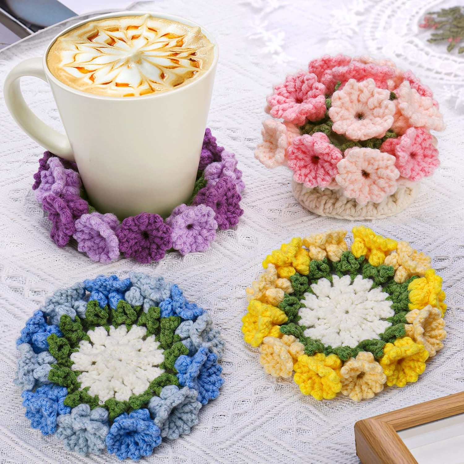 A mug of coffee on a purple handmade coaster from the Crochet Flower Coaster Kit 4pcs, with three other colorful floral coasters arranged on a white tablecloth.